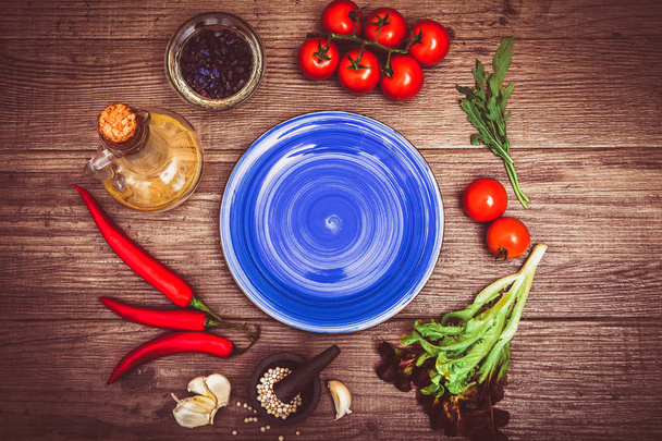 Fresh tomatoes, chili pepper and other spices and herbs around modern blue plate in the center of wooden table and cloth napkin. Top view. Blank place for your text. Close-up. - Photo, Image