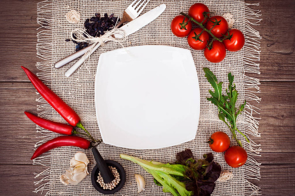 Fresh tomatoes, chili pepper and other spices and herbs around modern white square plate in the center of wooden table and cloth napkin. Top view. Blank place for your text. Close-up. - Foto, Imagen