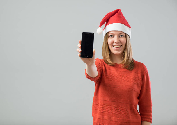 Young  woman with Santa hat showing smartphone - Foto, afbeelding