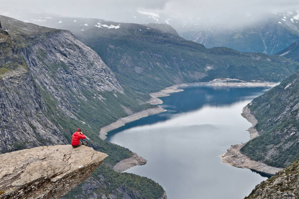 Hombre con cámara sentado en la roca Trolltunga (Troll 's Tongue rock) y hace la foto con el paisaje montañoso noruego
 - Foto, Imagen