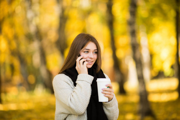 Tyttö puhuu puhelimessa kupin kahvia aurinkoisessa keltaisessa puistossa
 - Valokuva, kuva