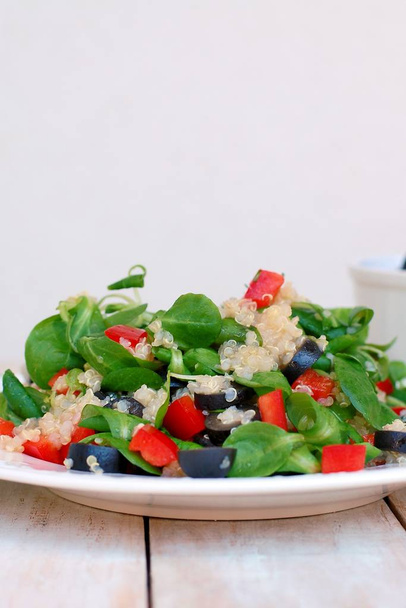 Fresh healthy vegan salad with quinoa, corn salad, black olives, red pepper and olive oil on white plate cloth on white wooden background - Photo, Image