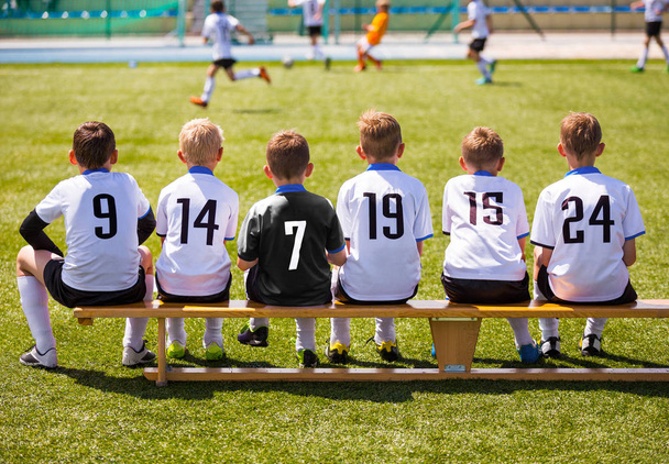 Ποδοσφαιριστές στον αγώνα παιχνίδι. Ομάδα νέων ποδοσφαίρου κάθεται στο ξύλινο πάγκο. Αγώνα ποδοσφαίρου για παιδιά. Μικρά αγόρια παίζουν αγώνα ποδοσφαίρου τουρνουά. Νεολαία Ποδόσφαιρο Club ποδοσφαιριστές - Φωτογραφία, εικόνα