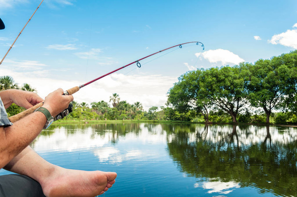 Man catching a fish with the rod enverged, holding a rod with his hands on a lake with a beautiful nature around. - Photo, Image