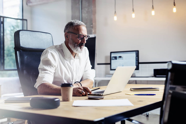 Adult professional businessman wearing a classic glasses and working at the wood table in modern coworking studio.Stylish bearded middle age man using laptop on workplace. Horizontal,blurred. - Foto, Bild