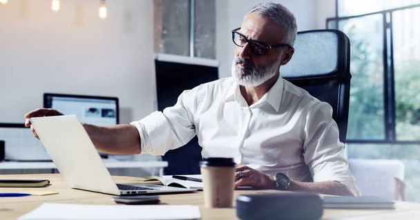 Middle age top manager wearing a classic glasses and working at the wood table in modern interior design office.Stylish bearded businessman using laptop on workplace. Horizontal wide,blurred. - Foto, Bild