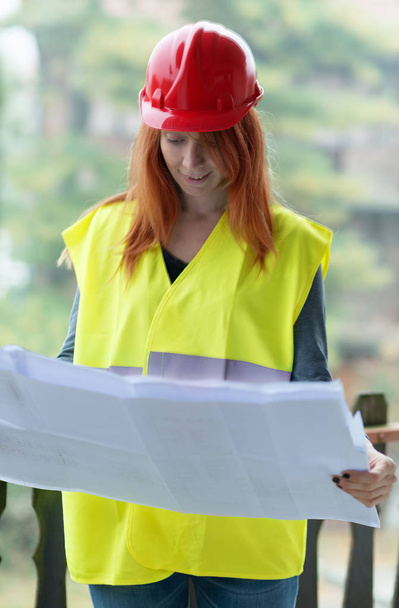 Mladá zrzka žena architekt červenou ochrannou přilbu a studovat plán výstavby.  - Fotografie, Obrázek