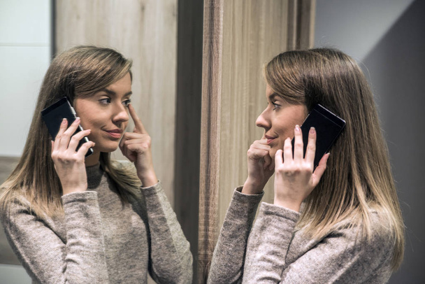 Porträt einer schönen jungen Frau am Telefon. Porträt einer glücklichen Frau, die ihr Spiegelbild im Spiegel betrachtet und telefoniert - Foto, Bild