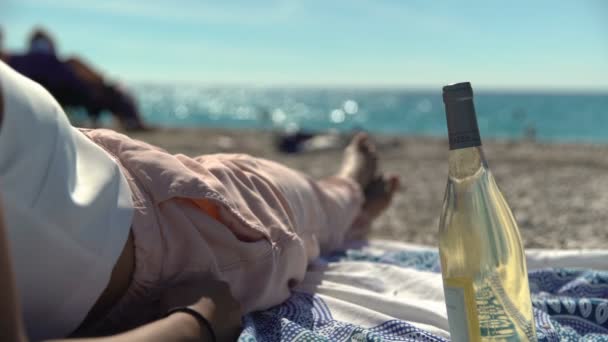 Femme avec du vin sur la Côte d'Azur. Une femme frissonne et se détend avec une bouteille de vin sur une plage tout en agitant ses orteils dans le sud de la France alors que les vagues s'écrasent en arrière-plan
. - Séquence, vidéo