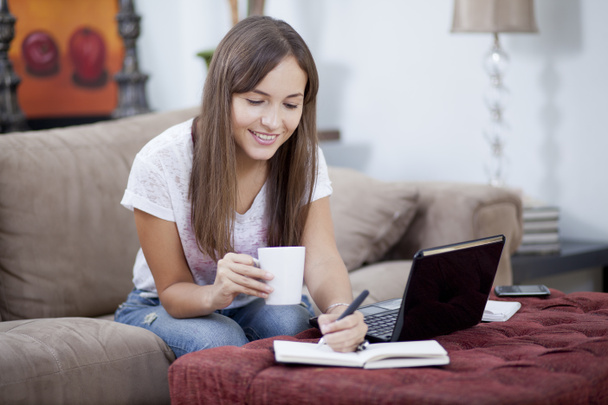 Nuori kaunis hymyilevä nainen istuu kannettavan tietokoneen ääressä ja tekee muistiinpanoja päiväkirjaansa ja ottaa kupin teetä.
 - Valokuva, kuva