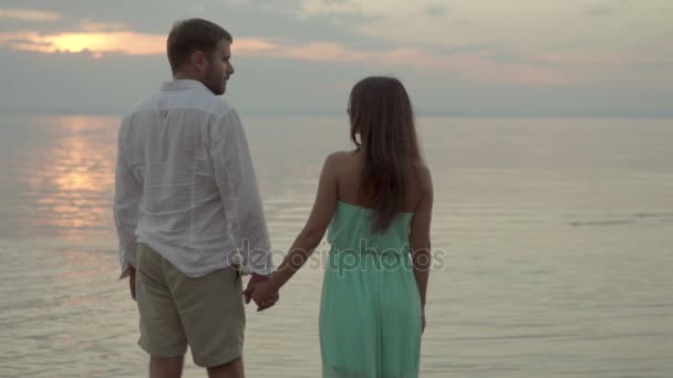 Feliz pareja joven en la playa al atardecer se toman de la mano y se miran el uno al otro
 - Metraje, vídeo