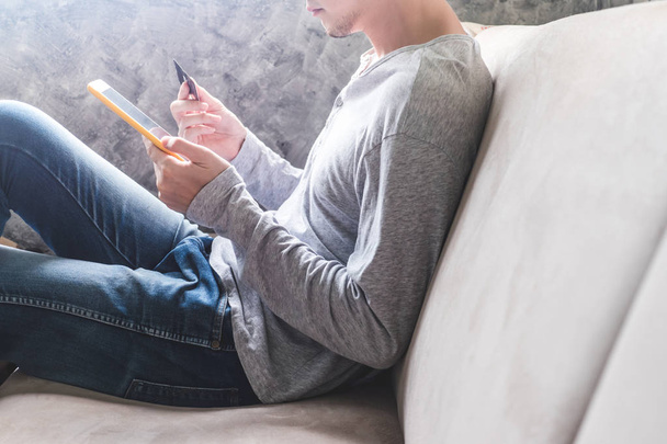 Casual young man using tablet and holding credit card on sofa at home. Online shopping concept. - Zdjęcie, obraz