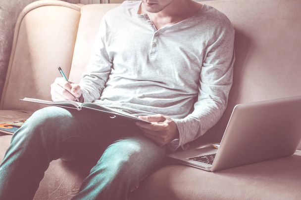 Giovane uomo casual che scrive alcuni dati in notebook con laptop sul divano di casa. tono vintage
 - Foto, immagini
