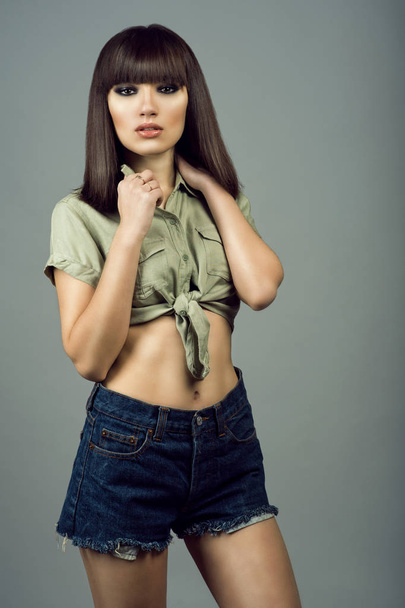 Portrait of young gorgeous dark-haired girl with provocative make up wearing high waisted dark blue jeans shorts and khaki tied up shirt with short sleeves. - 写真・画像