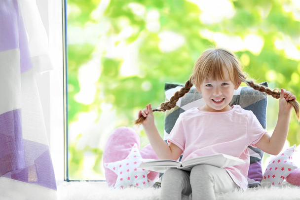 Beautiful little girl with book on windowsill - Photo, Image