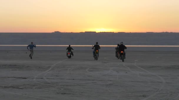 Bikers ride into sunset on lake, river, sea, bay. Man flees, falls and tumbles. - Footage, Video