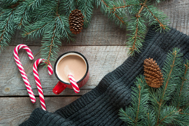 Mug hot coffee with milk, red candy cane on the wooden background with cone, branch of christmas tree and grey scarf. New Year. Holiday card. Rustic style. Top view and copy space. - 写真・画像