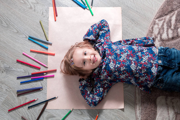 Dziecko leżące na podłodze papier patrząc na kamery w pobliżu kredki. Mała dziewczynka, malarstwo, rysunek. Widok z góry. Koncepcja kreatywność. - Zdjęcie, obraz