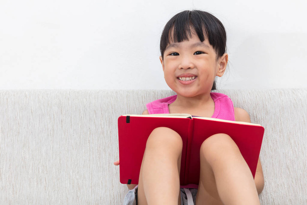 Happy Asian Chinese little girl sitting on sofa with book - Photo, Image