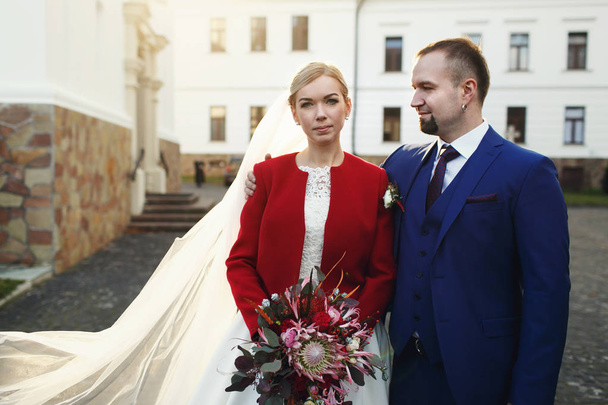Handsome groom in blue suit pose with a bride in red jacket  - Фото, изображение