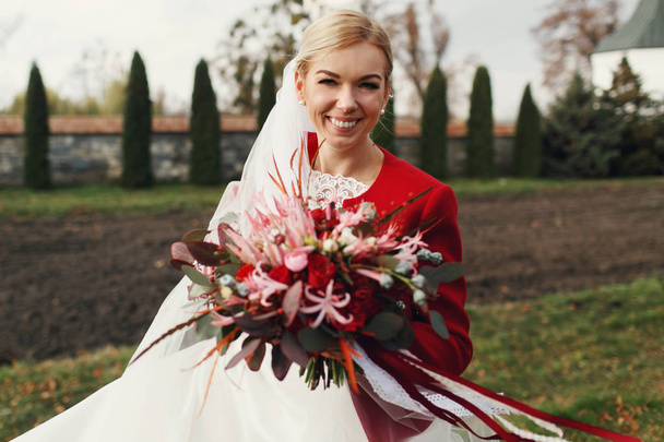 Beautiful blonde bride smiles posing in a red jacket in park  - Photo, image