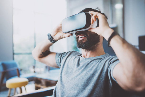 Bearded man wearing virtual reality goggles in modern coworking studio. Smartphone using with VR headset. Horizontal, blurred. - Φωτογραφία, εικόνα