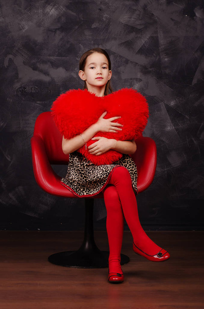 Pretty little girl wearing beautiful dress sitting in red armchair. She is holding plush heart in hands. Studio shot. - 写真・画像