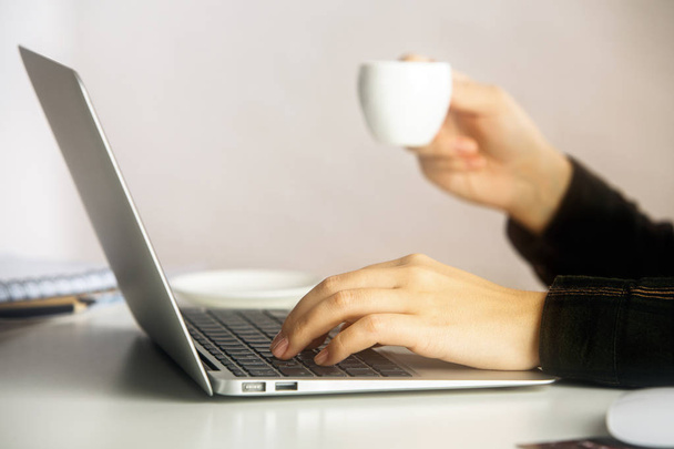 Side view of girl's hands holding coffee cup and typing on laptop keyboard placed on white office desktop. Communication concept - Zdjęcie, obraz