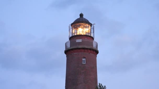 Shinning old lighthouse above pine forest before sunset. Tower illuminated with strong warning light. Lighthouse built from red bricks, gallery with iron handrail around glass cover of spotlight.  - Footage, Video