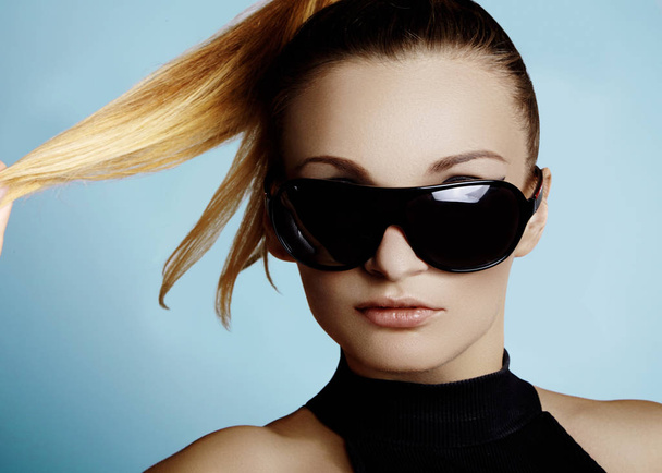Beautiful young woman with black fashion sunglasses and glamour pinytail hairstyle an lue background. Trendy look for lady. Eye wear style - Foto, imagen