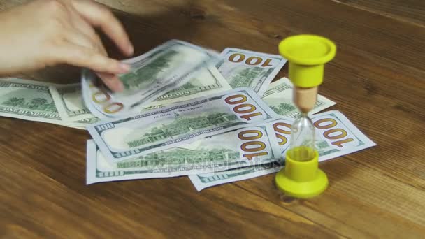 Billetes de dólar cayendo sobre una mesa de madera y reloj de arena
 - Metraje, vídeo