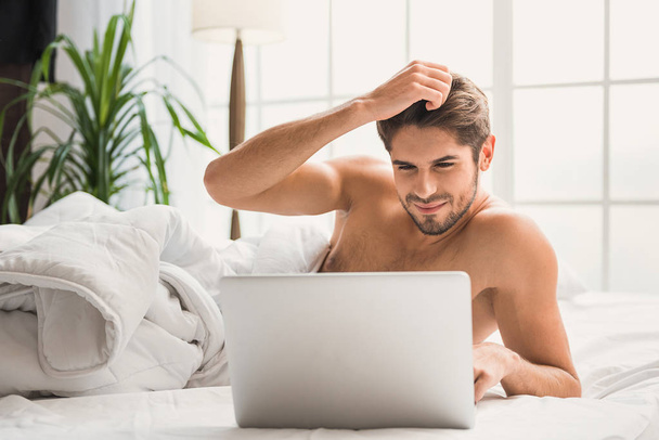 Carefree guy using computer in bedroom - Фото, изображение