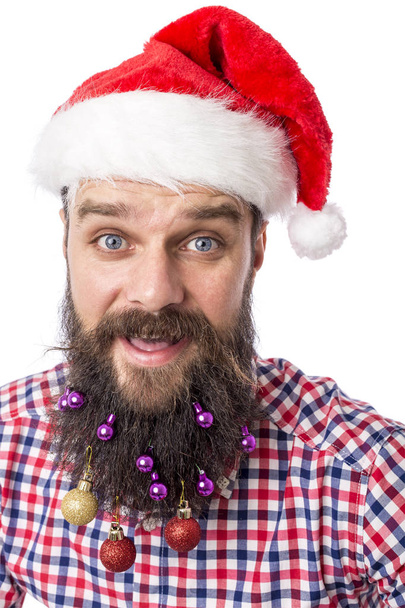 Closeup portrait of a funny man with colorful Christmas balls in his beard and santa red hat over white background - Photo, Image