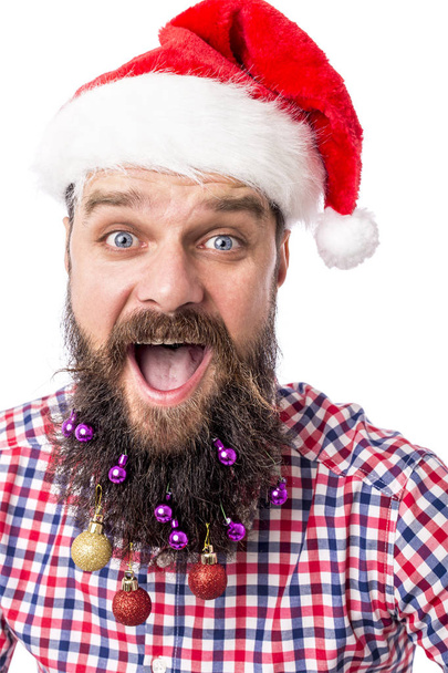 Closeup portrait of a funny man with Christmas balls in his beard and santa red hat over white background. - Фото, зображення