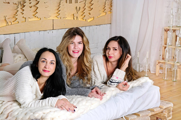 Три дівчини лежать на ліжку в затишних светрах і посміхаються
 - Фото, зображення