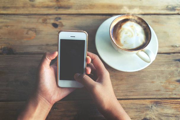 τα χέρια της γυναίκας κρατώντας το κινητό έξυπνο τηλέφωνο με φλιτζάνι ζεστό καφέ στο καφέ shop - Φωτογραφία, εικόνα