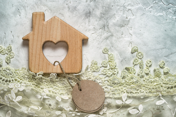 Wooden toy house on a cement background with lace grass and tied blank round paper tag. Copy space for your inscription. - Photo, Image