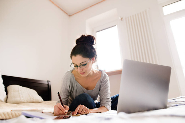 Beautiful young woman sitting on bed, working. Home office. - Фото, изображение