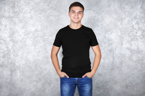 Handsome young man in blank black t-shirt standing against grey textured wall - Фото, зображення