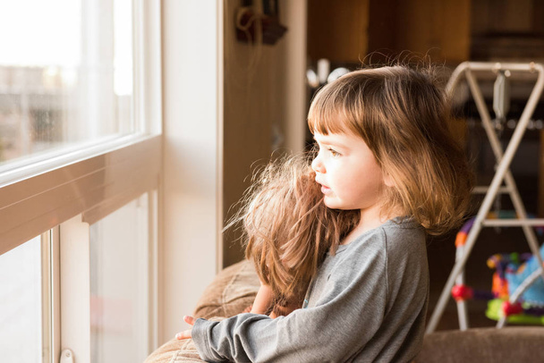 linda chica joven morena juega con la muñeca mientras usa una camisa gris y de pie cerca de la ventana
 - Foto, Imagen