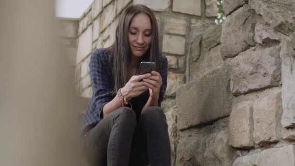美しい若い女性は、幸せな時間のレンガの壁の内部に屋外で携帯電話を使用します。 - 映像、動画