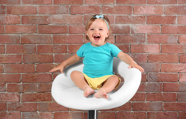 Baby girl in color dress sitting on a bar chair on a brick wall background - Fotografie, Obrázek