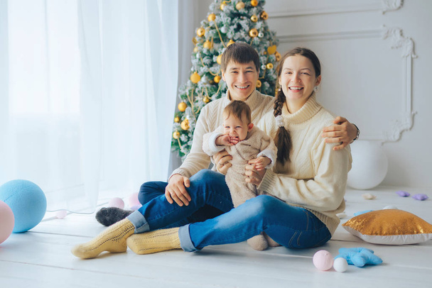 Jonge ouders zitten onder de kerstboom met Malenkov schattige dochter in haar armen, glimlachen en kijken naar het frame - Foto, afbeelding