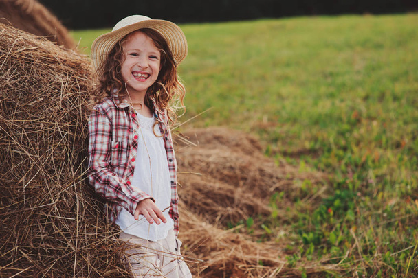 gelukkig kind meisje in land stijl plaid shirt en Pet ontspannen op zomer veld met hooi stapels - Foto, afbeelding