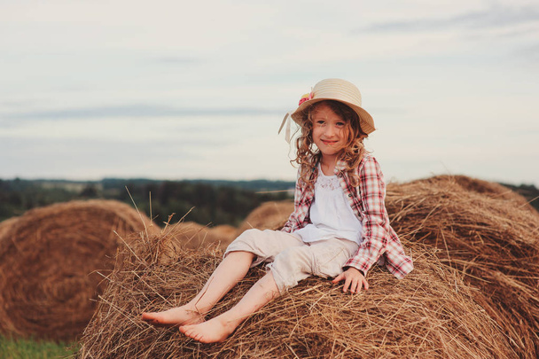 happy child girl in country style plaid shirt and hat relaxing on summer field with hay stacks - Zdjęcie, obraz