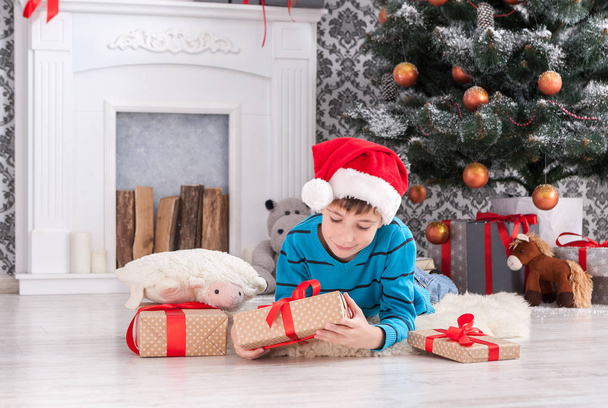 Симпатичный мальчик в шляпе Санты разворачивает рождественские подарки
 - Фото, изображение