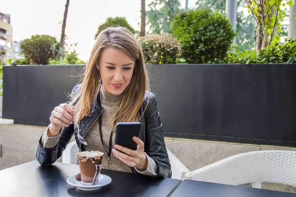 Κορίτσι γραπτών μηνυμάτων για το έξυπνο τηλέφωνο σε ένα εστιατόριο με βεράντα με μια αόριστες φόντο, νεολαία γυναίκα Coffee Shop χρησιμοποιώντας έξυπνο τηλέφωνο έννοιας, γυναίκα χρησιμοποιώντας έξυπνο τηλέφωνο Coffee Shop έννοιας - Φωτογραφία, εικόνα