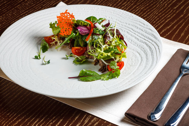 Vegetable salad menu for the restaurant in a large white plate on a white table cloth on a wooden table. Salad on the table with knife and fork. Svezhive vegetables. - 写真・画像