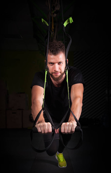 One young handsome brave curly bearded muscled man with a beard makes Total Resistance eXercises fitness trx straps working out in gym - Valokuva, kuva