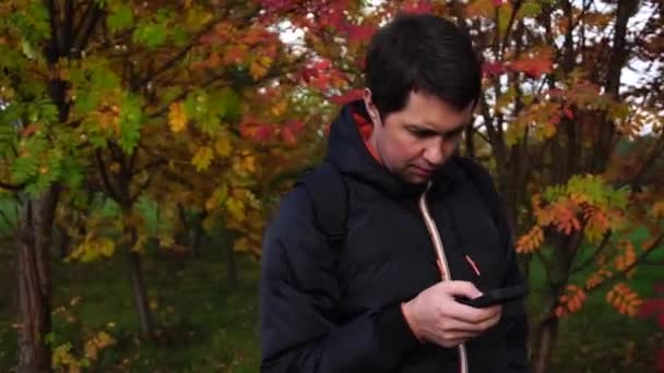 Fiatal férfi fekete kabát, az őszi parkban használ a smartphone - Felvétel, videó